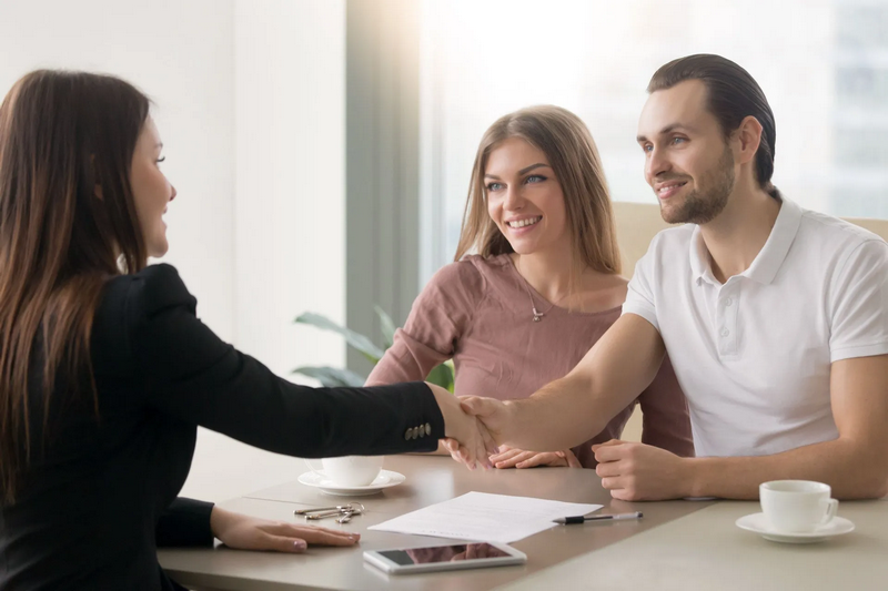 A couple is shaking hands with a businesswoman across a table in an office setting.