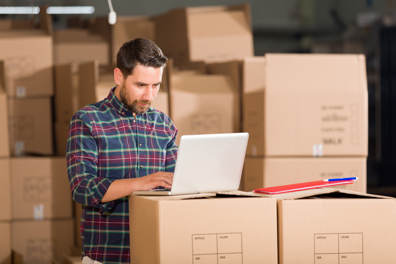 A man stands in a warehouse, using a laptop on top of cardboard boxes.