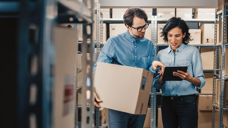 Two people in a warehouse checking inventory on a tablet while holding a cardboard box.