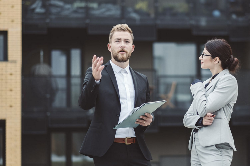 A man in a suit is speaking while holding a folder, and a woman in a suit listens with her hand on her chin