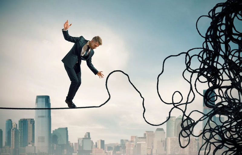 A man in a suit balances on a tightrope with a chaotic knot on one end, against a city skyline backdrop.